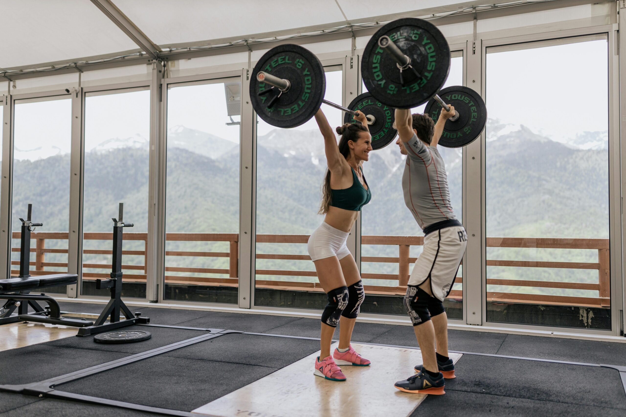 A couple lifting barbells together in a gym with scenic mountain views.